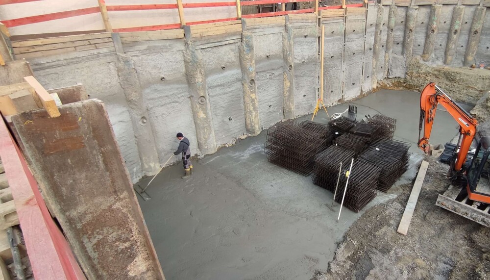 Bauarbeiter glättet Beton in einer großen Baugrube mit Stahlarmierungen und Bagger