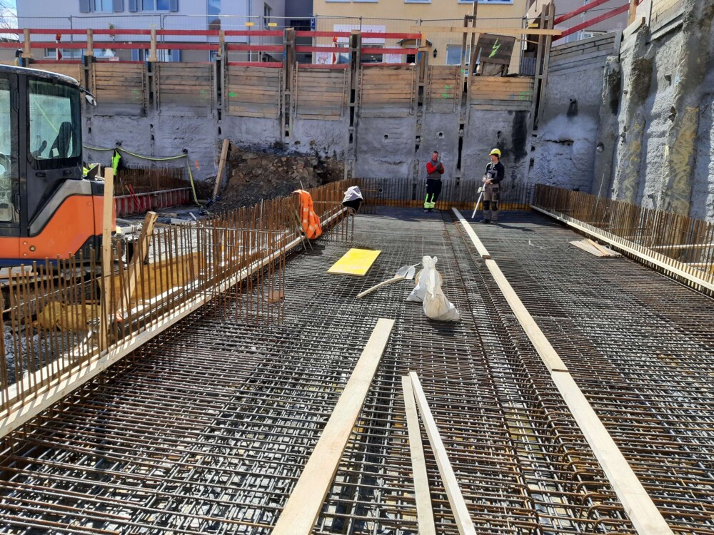 Baustelle mit Bewehrungseisen für Betonboden, zwei Bauarbeiter mit Schutzhelmen und Baugerät links