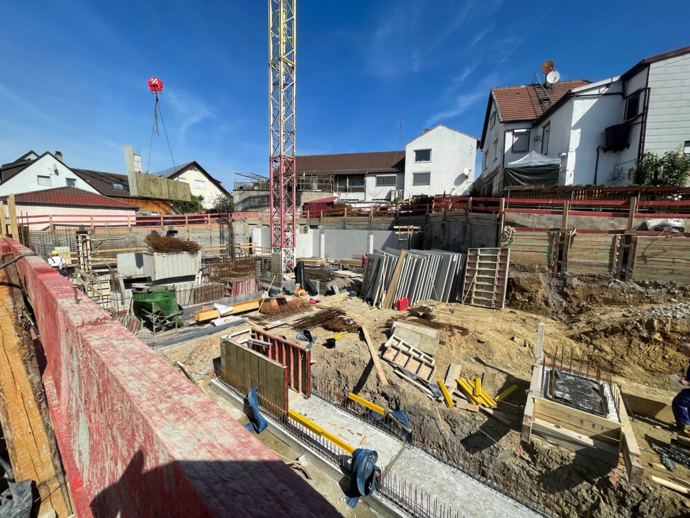 Baustelle mit Baugrube, Schalungen und Kran, umgeben von Wohnhäusern unter blauem Himmel