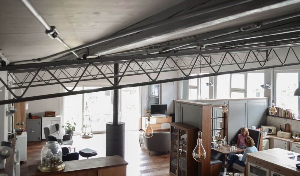 Innenansicht eines Loftgebäudes mit sichtbaren Stahlträgern, großen Fenstern und modern eingerichteten Wohn- und Arbeitsbereichen