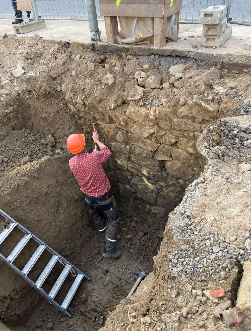 Archäologe mit orangefarbenem Helm und rotem Hemd untersucht eine freigelegte Stadtmauer in einer Ausgrabungsgrube