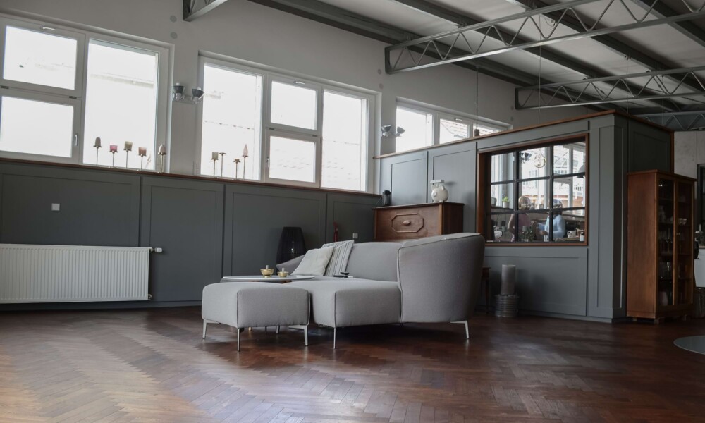 Großes, helles Wohnzimmer mit grauem Sofa, Holzboden und hohen Fenstern in einem Industriegebäude