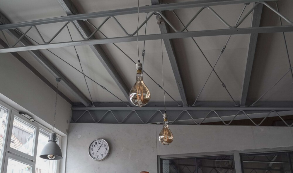 Loftgebäude mit sichtbaren Stahlträgern an der Decke, zwei hängende Glühbirnen und eine Wanduhr mit arabischen Zahlen